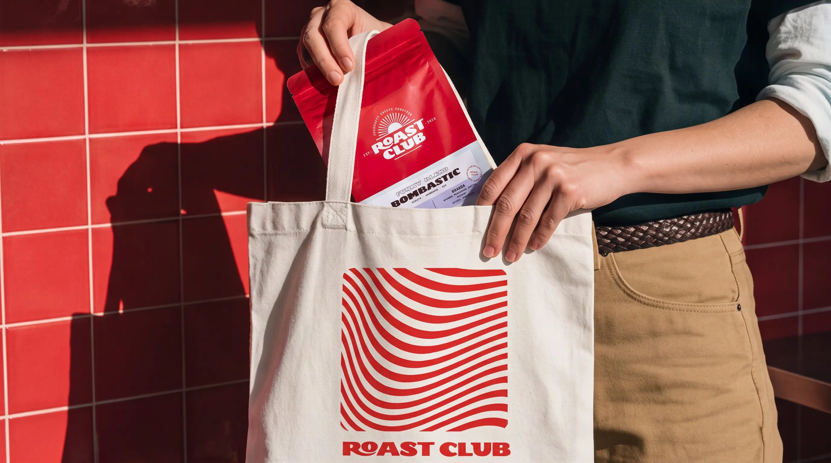 Mano guardando una bolsa de café Roast Club en una tote de la marca, sobre azulejo rojo